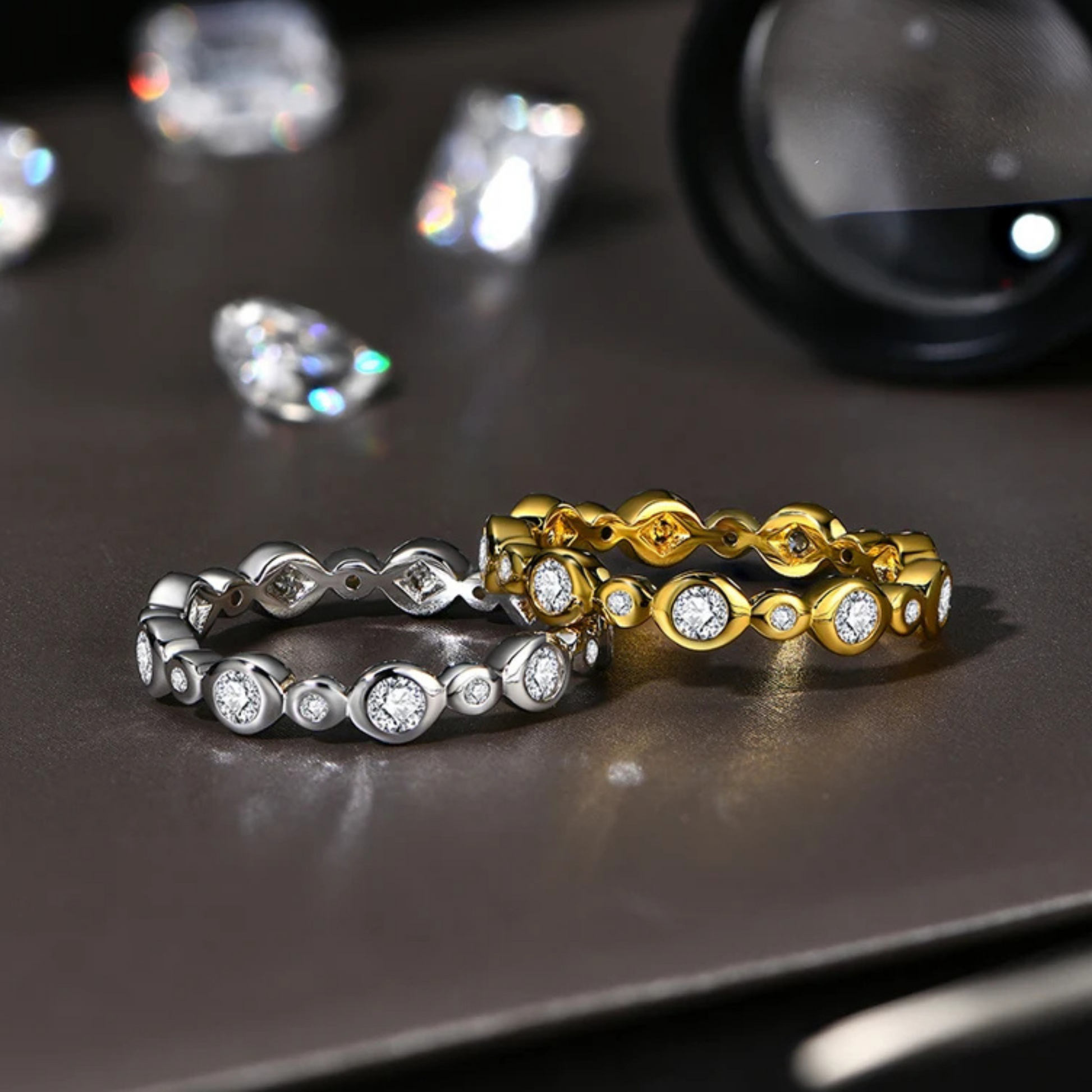 Two moissanite-studded rings, one silver and one gold, on a dark surface with loose diamonds.